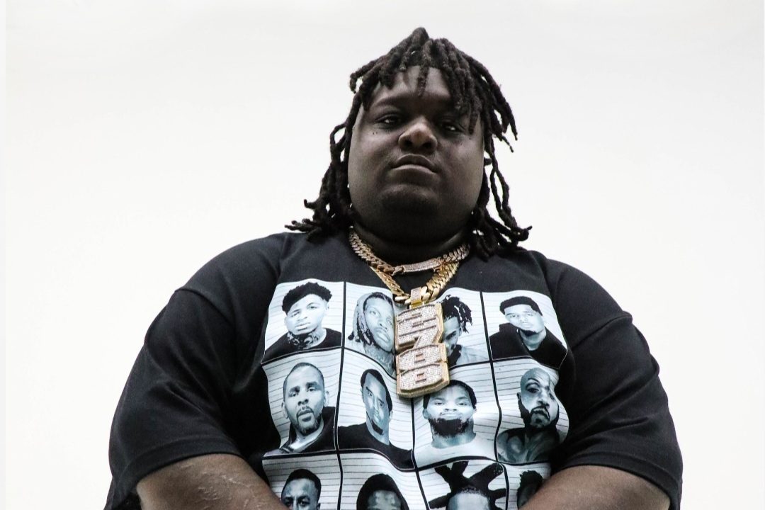 Dallas rapper HeadHuncho Amir wears a black shirt and gold chains standing in front of a white background looking straight to the camera.