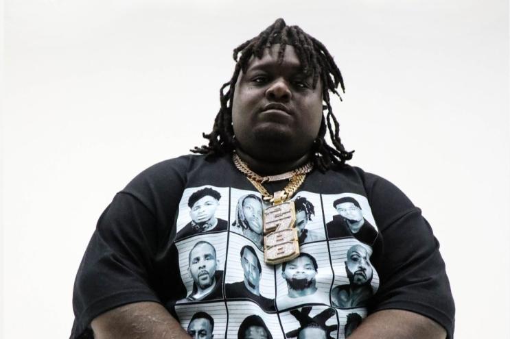 Dallas rapper HeadHuncho Amir wears a black shirt and gold chains standing in front of a white background looking straight to the camera.