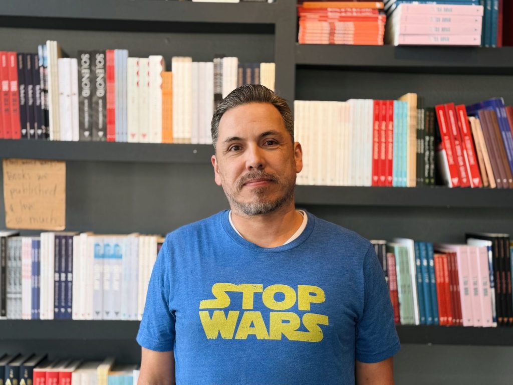 Poet Joaquín Zihuatanejo stands before a wall of books at Deep Vellum,