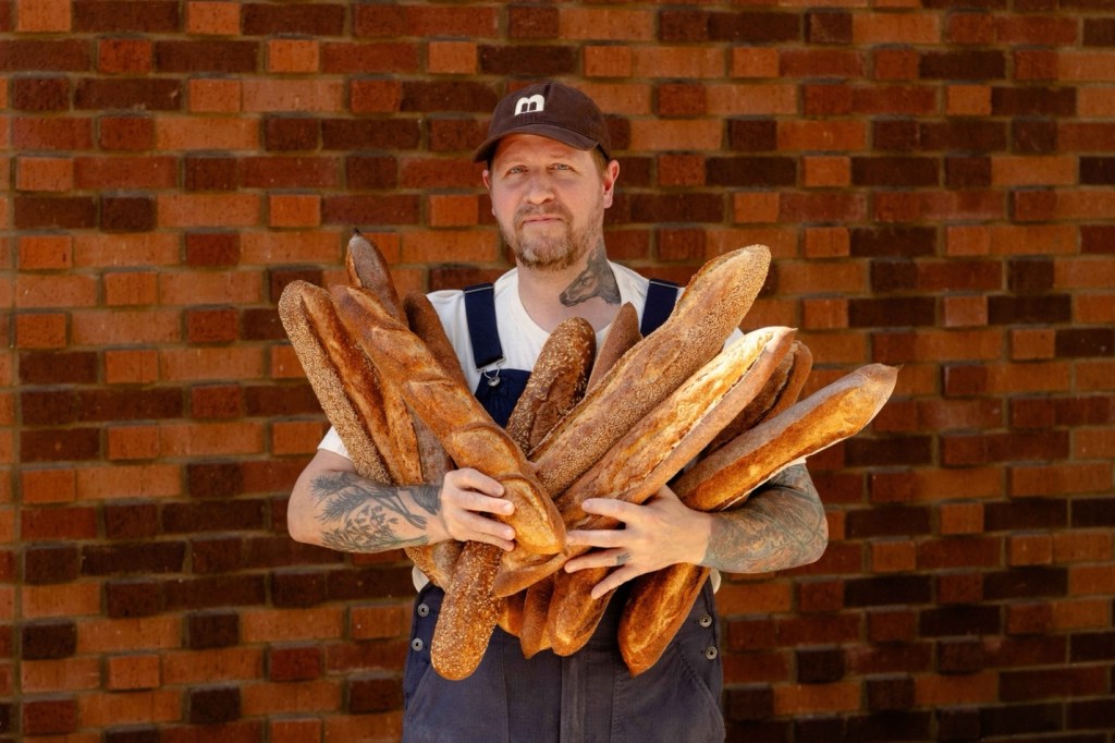 A photo of The Bread Club's Baking Director Peter Edris, holding an armful of baguettes infront of a brick wall.