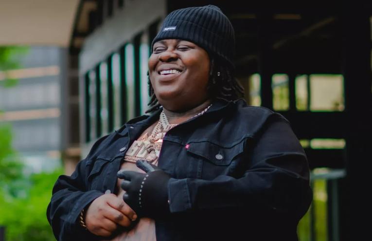 Rapper BigXthaPlug smiling in front of building with shirt unbuttoned wearing a beanie